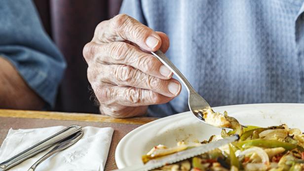 Eine ältere Hand hält eine Gabel über einem Teller mit gebratenem Gemüse und Fleisch.